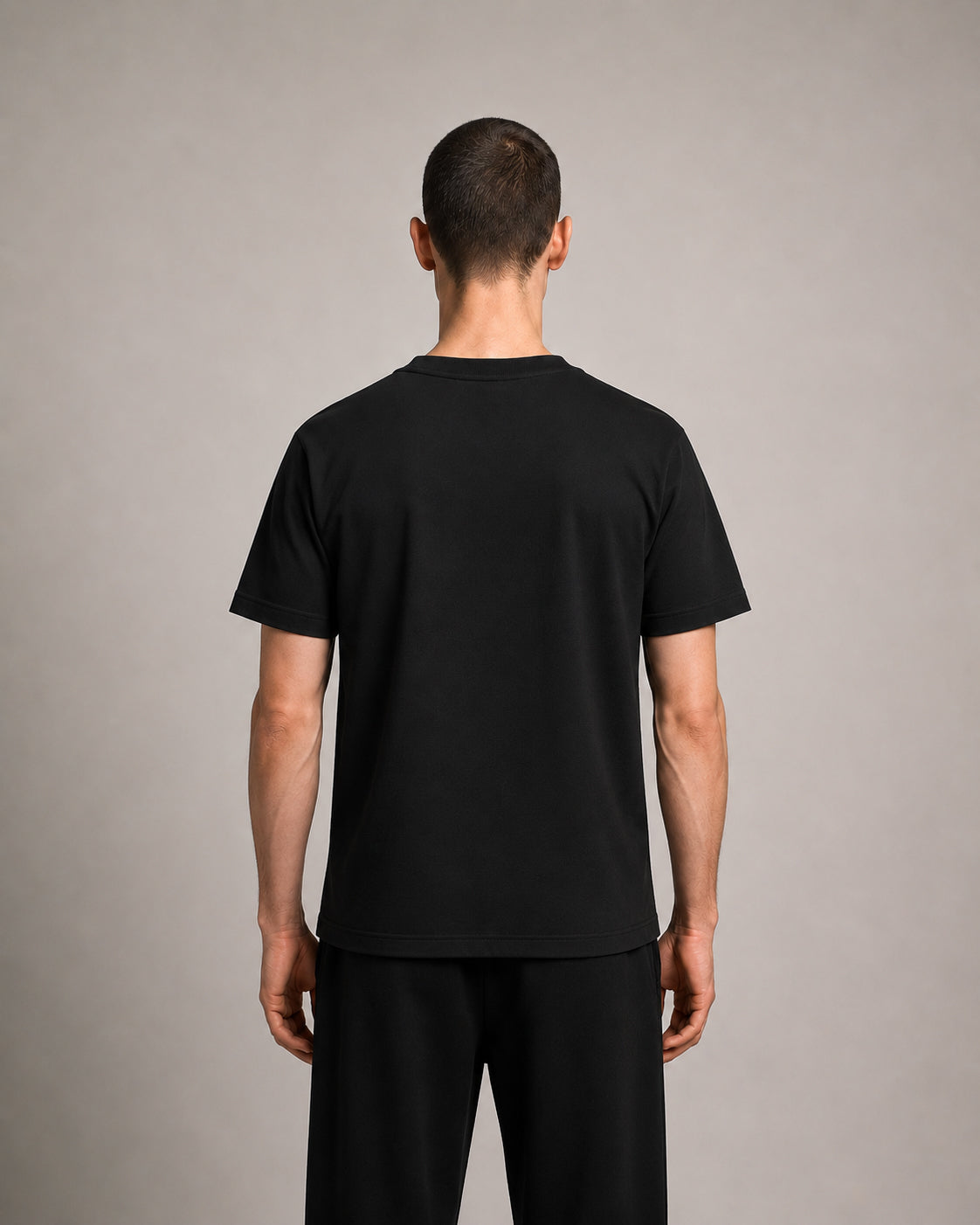 A man viewed from the back wearing a plain black short-sleeve t-shirt and black pants against a neutral gray studio background.