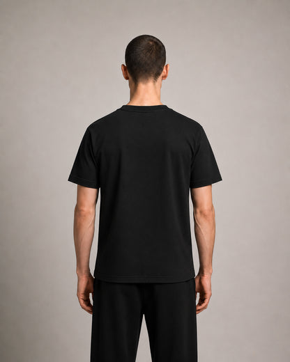 A man viewed from the back wearing a plain black short-sleeve t-shirt and black pants against a neutral gray studio background.