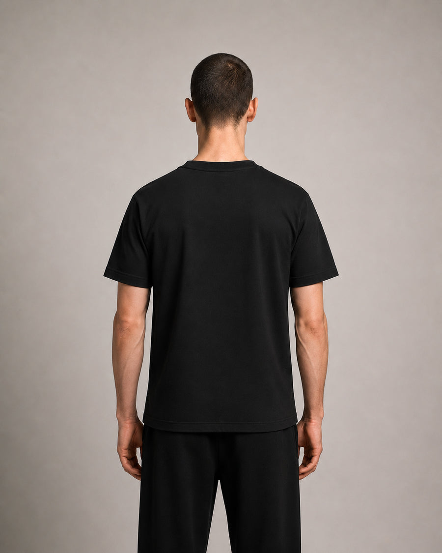 A man viewed from the back wearing a plain black short-sleeve t-shirt and black pants against a neutral gray studio background.
