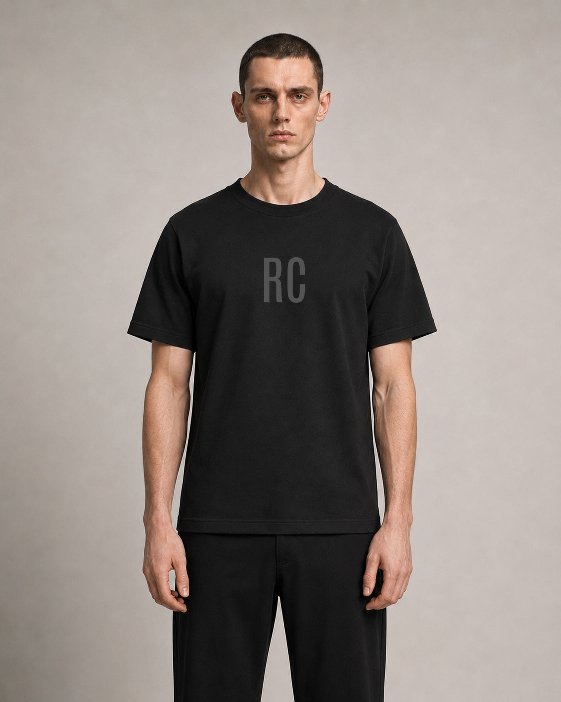 A man wearing a black short-sleeve crewneck t-shirt with a subtle gray 'RC' logo on the chest, paired with black pants, standing against a neutral gray studio background.