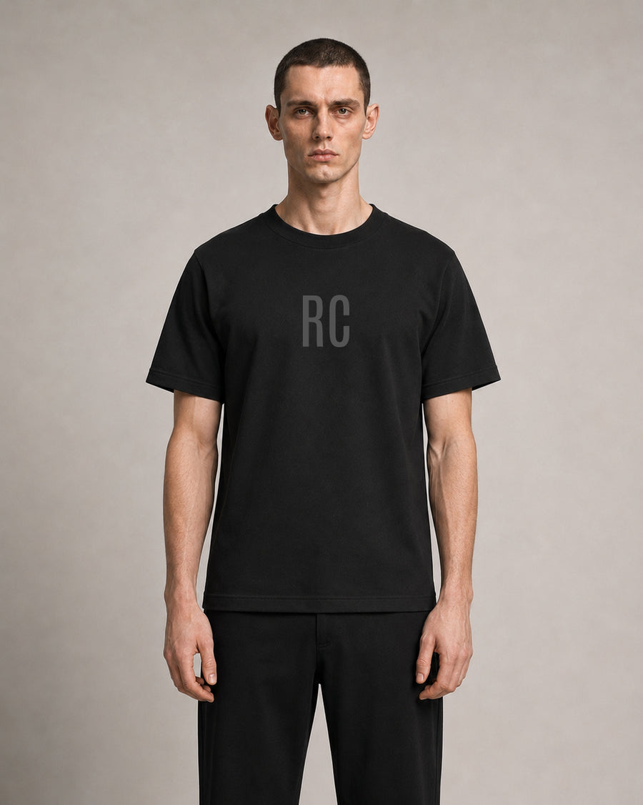 A man wearing a black short-sleeve crewneck t-shirt with a subtle gray 'RC' logo on the chest, paired with black pants, standing against a neutral gray studio background.