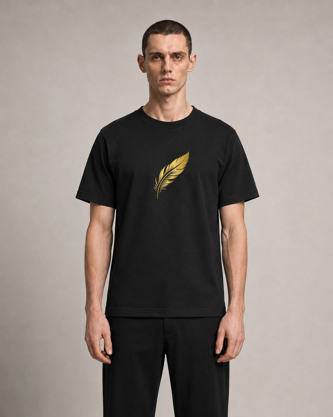 A man wearing a black short-sleeve crewneck t-shirt with a metallic gold feather graphic on the chest, paired with black trousers, standing against a neutral gray studio background.