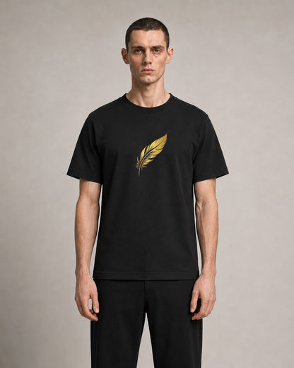 A man wearing a black short-sleeve crewneck t-shirt with a metallic gold feather graphic on the chest, paired with black trousers, standing against a neutral gray studio background.